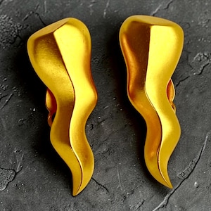 May include: Two gold-colored, abstract-shaped earrings. Each earring has a curved, horn-like design with a smooth, reflective surface. The earrings are displayed against a dark, textured background.