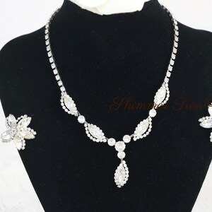 May include: A sparkling silver-toned necklace and matching earrings set. The necklace features a series of clear, round and marquise-shaped crystals. The earrings are flower-shaped with multiple clear crystals. The jewelry is displayed on a black mannequin.