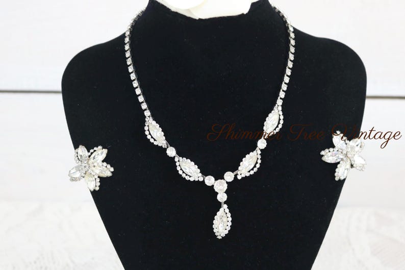 May include: A sparkling silver-toned necklace and matching earrings set. The necklace features a series of clear, round and marquise-shaped crystals. The earrings are flower-shaped with multiple clear crystals. The jewelry is displayed on a black mannequin.