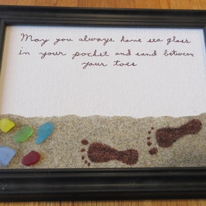May include: A framed piece of art with a handwritten message that reads "May you always have sea glass in your pocket and sand between your toes". The art piece features a small beach scene with brown footprints and colorful sea glass.