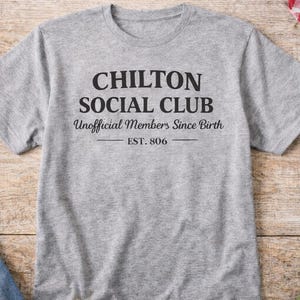 May include: A heather gray t-shirt with the text "CHILTON SOCIAL CLUB Unofficial Members Since Birth EST. 806" in black. The shirt is on a wooden surface, with a glass of water and jeans visible.