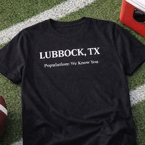 May include: Black t-shirt with the text "LUBBOCK, TX" and "Population: We Know You" in white. A brown football and a red cooler with sunglasses are also visible on a green field with white lines.