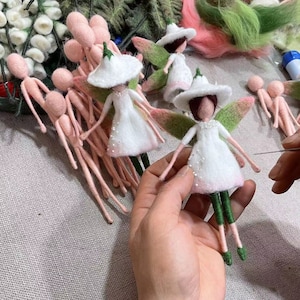 Puede incluir: Primer plano de una mano sosteniendo una muñeca de hada de fieltro con alas verdes, un vestido blanco y un sombrero verde. La muñeca está siendo cosida con una aguja e hilo. Otras muñecas de fieltro son visibles en el fondo.