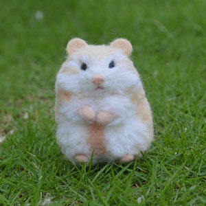 May include: A small, white and light brown hamster figurine with a fluffy texture. The hamster is standing on its hind legs with its paws together in front of its chest. The figurine is sitting on a bed of green grass.