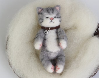 May include: A gray and white needle felted cat with a brown collar and a silver bell, lying in a white fluffy nest.