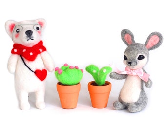 Op de afbeelding: Twee vilten dierenfiguren, een witte hond met een bril en een rode sjaal, en een grijs konijn met een roze strik, staan naast twee kleine groene planten in potten.