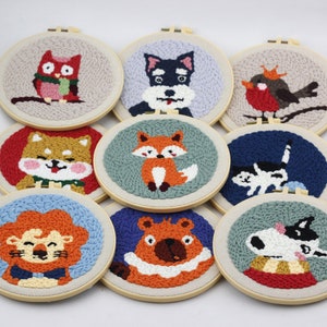 Könnte beinhalten: Neun runde Stickrahmen mit verschiedenen Tiermotiven. Die Motive umfassen einen Fuchs, eine Katze, einen Hund, einen Löwen, einen Bären, eine Eule, einen Rotkehlchen und einen Shiba Inu. Die Rahmen sind aus Holz und haben eine natürliche Oberfläche.