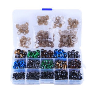 May include: A clear plastic storage box filled with assorted plastic safety eyes for crafts. The box has 12 compartments, each containing a different color and size of eyes. The eyes are black, brown, gold, green, and blue.