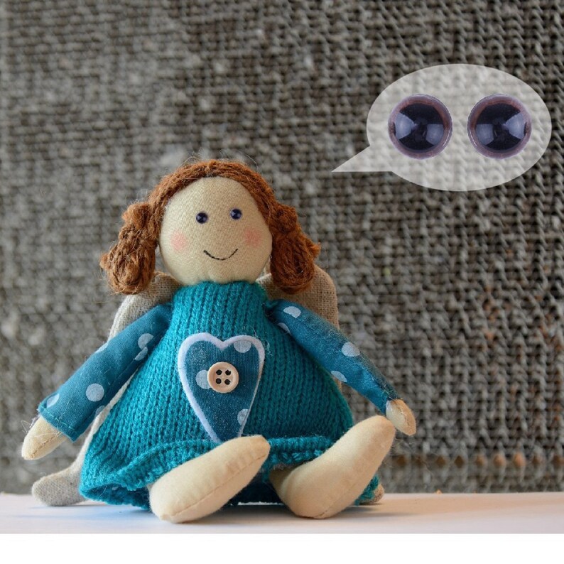May include: A handmade blue and white angel doll with brown hair and a heart-shaped design on her chest. The doll is sitting on a white surface and has a speech bubble above her head with two brown eyes.