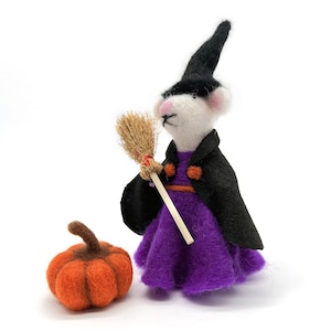 Puede incluir: Un ratón de fieltro blanco vestido de bruja con un sombrero negro, un vestido morado y una capa negra. El ratón sostiene una escoba marrón y está de pie junto a una calabaza de fieltro naranja.