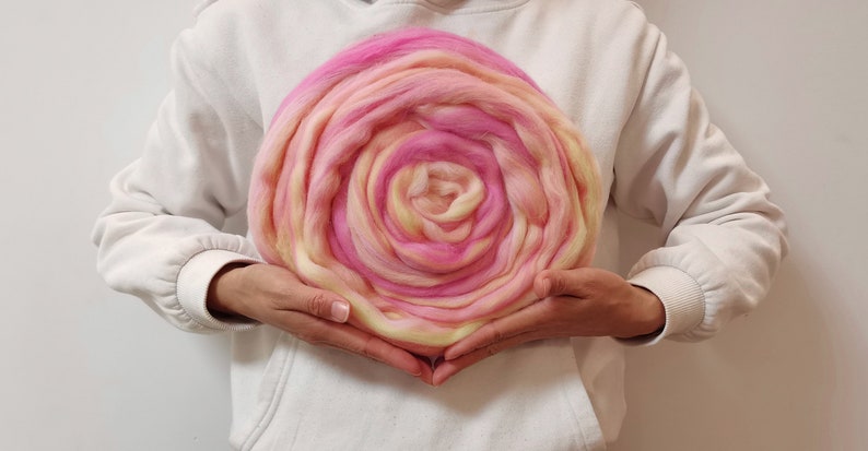 May include: A large, circular, pink and yellow wool roving, resembling a rose, held in the hands of a person.