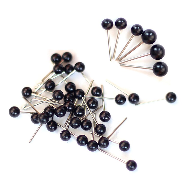 May include: A pile of black and silver push pins with round heads. The pins are scattered on a white surface.