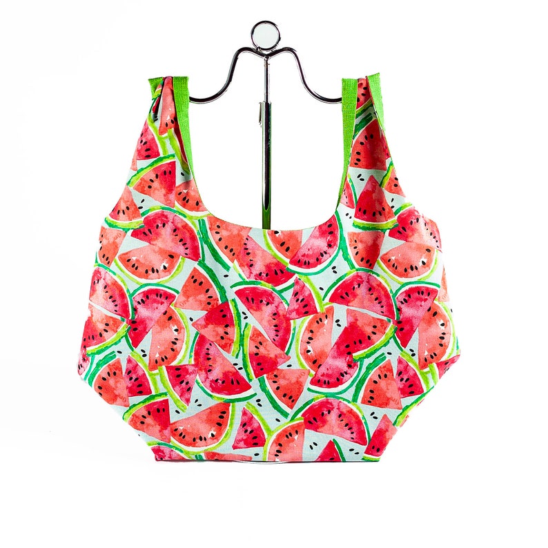 May include: A reusable shopping bag with a watermelon print. The bag is made of a lightweight fabric and has a green trim. It is folded and displayed on a metal stand.