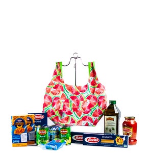 May include: A watermelon-patterned reusable shopping bag filled with grocery items, including pasta, canned goods, olive oil, and tomato sauce. The bag is hanging from a metal hook.