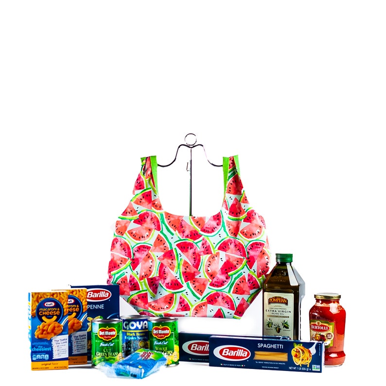 May include: A watermelon-patterned reusable shopping bag filled with grocery items, including pasta, canned goods, olive oil, and tomato sauce. The bag is hanging from a metal hook.