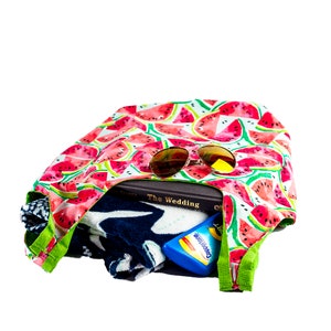 May include: A watermelon print tote bag filled with a book, a towel, sunglasses, and sunscreen. The book is titled "The Wedding" and the sunscreen bottle says "Coppertone".