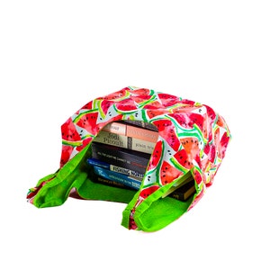 May include: A green and white reusable tote bag with a watermelon print. The bag is filled with a stack of paperback books. The titles of the books are visible: "The Hour", "Jodi Picoult", "Kristin Hannah", "Plain Truth", "All the Light We Cannot See", "Anthony Doerr", "Fishing North", "A Field Guide to the Birds", "Nicholas Sparks".