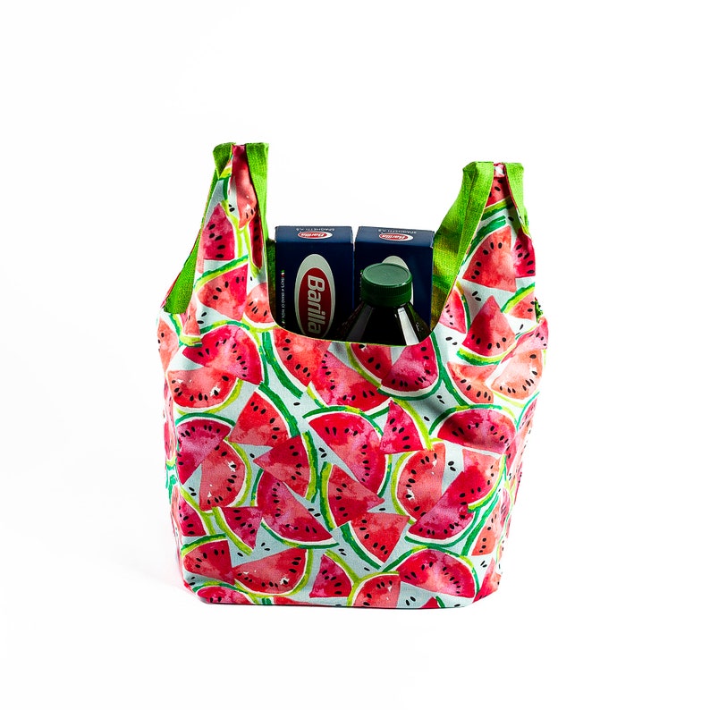 May include: A reusable shopping bag with a watermelon print. The bag is green and white with red watermelon slices. The bag is filled with two boxes of Barilla pasta and a green glass bottle.