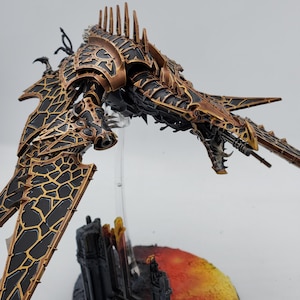 May include: A detailed, hand-painted miniature of a black and gold spaceship with a dragon-like head and wings. The spaceship is posed in mid-flight, with its wings spread wide. The miniature is set on a base with a black and orange cityscape.