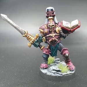 May include: A painted miniature figure of a warrior holding a sword and a book. The figure is wearing a red and gold armor with a blue and green cape. The figure is standing on a gray base with green grass.