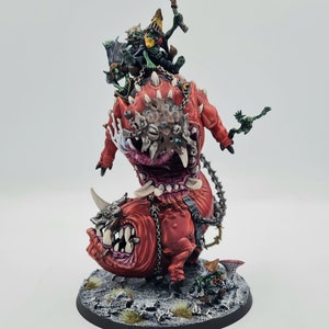 May include: A red and green miniature wargaming figure of a monstrous beast with a smaller beast attached to its back. The larger beast has a gaping maw with sharp teeth and a spiked collar. The smaller beast has a similar mouth and is being ridden by a green goblin-like figure.