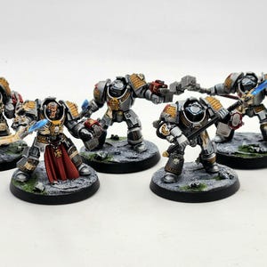 Puede incluir: Un grupo de cinco figuras en miniatura pintadas de gris y dorado, incluyendo una figura de líder con una capa roja, de pie sobre bases circulares negras. Las figuras sostienen espadas y otras armas.