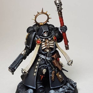 May include: A detailed miniature figure of a warrior in black armor, holding a staff with a skull-shaped top. The figure has a skeletal chest design, a halo, and is standing on a base with skulls.