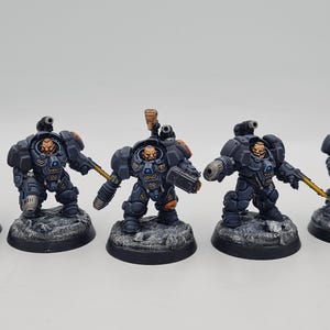 Könnte beinhalten: Fünf blau und grau bemalte Space Marine Miniaturen mit gelben Akzenten. Die Figuren halten Waffen und sind in Kampfstellung.