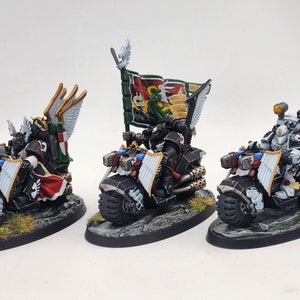 Może przedstawiać: Trzy miniaturowe figurki jeżdżące na motocyklach, pomalowane na czarno, biało i czerwono z złotymi akcentami. Każda figurka ma na sobie hełm i skrzydła na plecach. Figurki trzymają broń i jeżdżą na czarnych motocyklach z białymi kołami. Motocykle mają różne detale, w tym rury, światła i flagę.