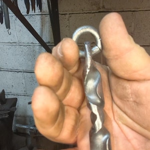 Pode incluir: Um gancho de metal forjado à mão com um design torcido e um laço circular no topo. O gancho é prateado e parece ser feito de ferro ou aço. O fundo mostra uma oficina.
