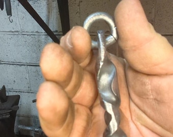 May include: A hand-forged metal hook with a twisted design and a circular loop at the top. The hook is silver-coloured and appears to be made of iron or steel. The background shows a workshop setting.