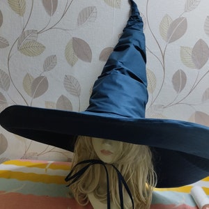 Puede incluir: Un gran sombrero de bruja de ala ancha con una corona alta y puntiaguda. El sombrero está hecho de una tela azul oscura y tiene una cinta negra atada alrededor de la base de la corona.