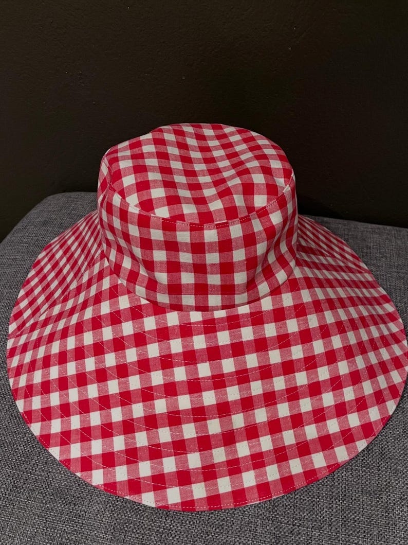 K&ouml;nnte beinhalten: Ein Sonnenhut mit breiter Krempe und rot-wei&szlig;em Vichy-Muster. Der Hut hat eine runde Krone und eine breite, kreisf&ouml;rmige Krempe. Der Stoff ist in einem klassischen Karomuster gehalten.