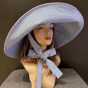 May include: A wide-brimmed sun hat with a blue and white gingham pattern. The hat features a large brim and a matching gingham bow tied under the chin. The hat is displayed on a mannequin head.