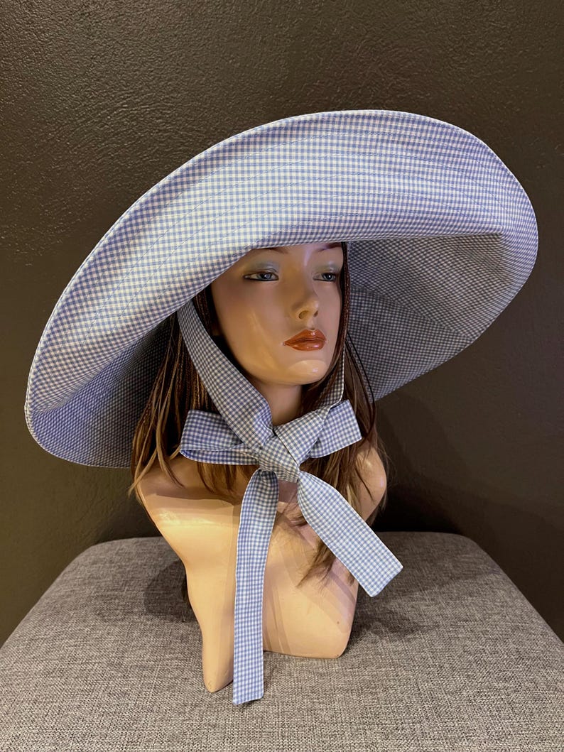 May include: A wide-brimmed sun hat with a blue and white gingham pattern. The hat features a large brim and a matching gingham bow tied under the chin. The hat is displayed on a mannequin head.