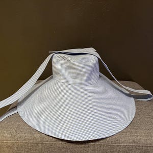 May include: A light blue and white gingham sun hat with a wide brim and long straps. The hat is sitting on a gray upholstered surface. The hat's design is a classic pattern, suitable for sun protection.