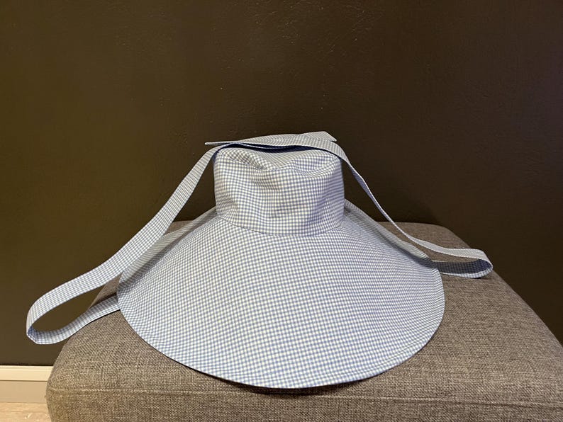 May include: A light blue and white gingham sun hat with a wide brim and long straps. The hat is sitting on a gray upholstered surface. The hat's design is a classic pattern, suitable for sun protection.