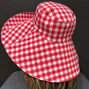 K&ouml;nnte beinhalten: Ein rot-wei&szlig; karierter Sonnenhut mit breiter Krempe. Der Hut ist aus Stoff gefertigt und hat ein klassisches Vichy-Muster. Der Hut ist f&uuml;r den Sonnenschutz konzipiert und eignet sich f&uuml;r Outdoor-Aktivit&auml;ten.