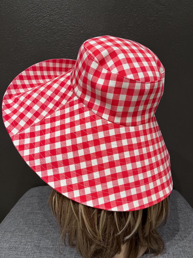 K&ouml;nnte beinhalten: Ein rot-wei&szlig; karierter Sonnenhut mit breiter Krempe. Der Hut ist aus Stoff gefertigt und hat ein klassisches Vichy-Muster. Der Hut ist f&uuml;r den Sonnenschutz konzipiert und eignet sich f&uuml;r Outdoor-Aktivit&auml;ten.
