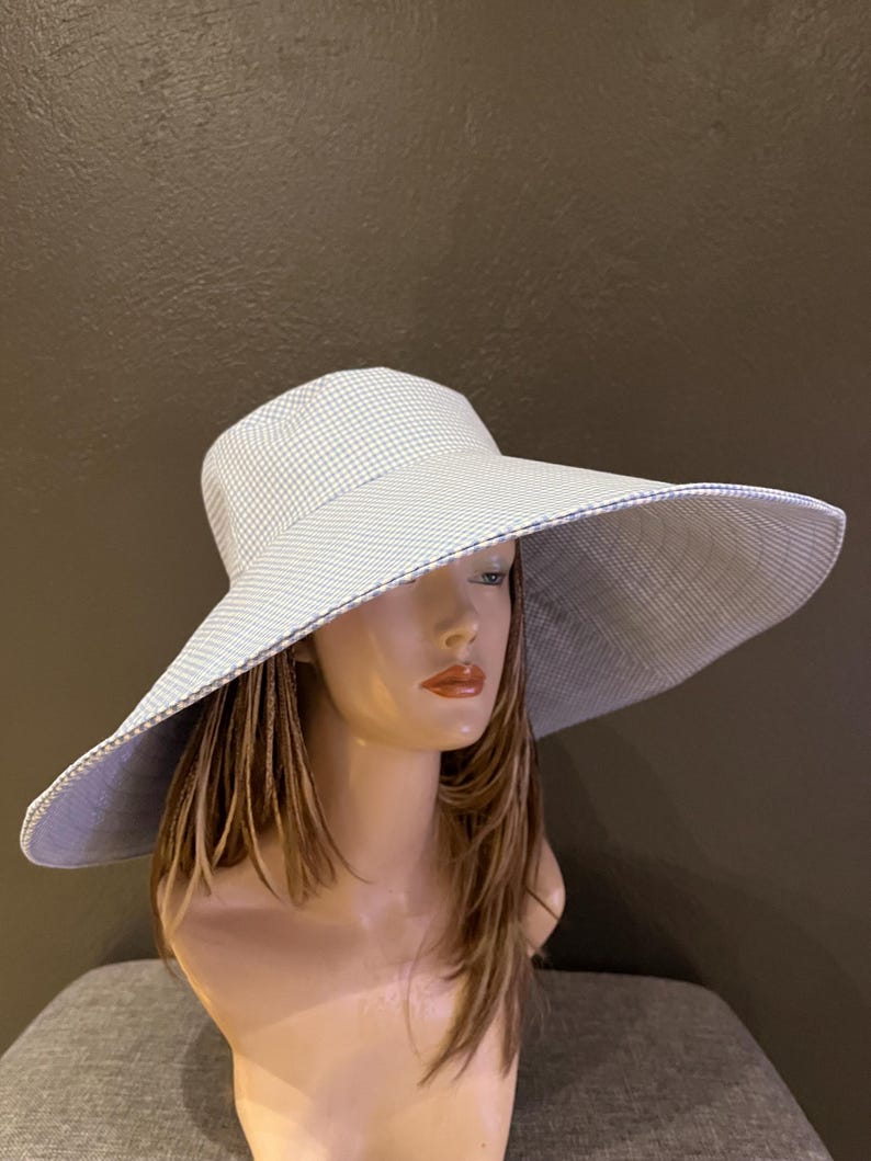 May include: A wide-brimmed sun hat with a light blue and white gingham pattern. The hat features a large brim and a matching interior lining. The hat is displayed on a mannequin head.