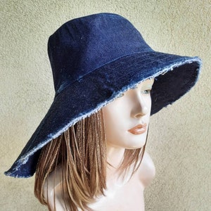Puede incluir: Un sombrero de cubo de denim de ala ancha con un borde deshilachado. El sombrero es azul oscuro y está hecho de una tela resistente.