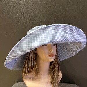 May include: A large, wide-brimmed hat with a blue and white gingham pattern. The hat is displayed on a mannequin head with long, light brown hair. The hat's brim is curved and provides ample shade.