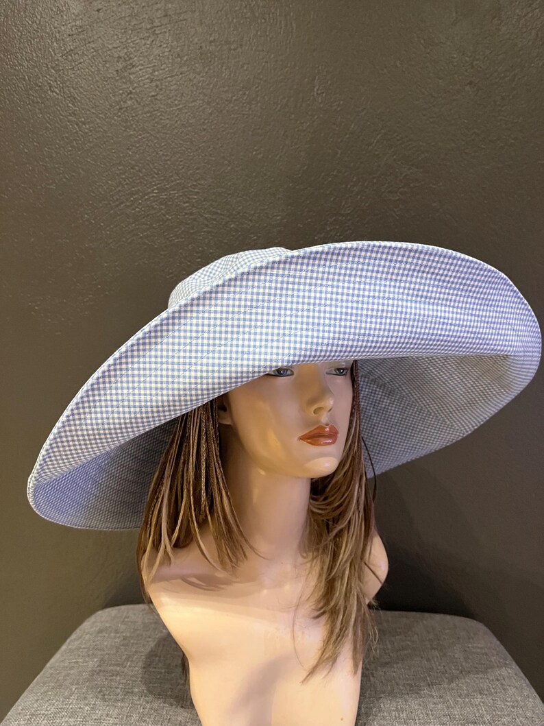 May include: A large, wide-brimmed hat with a blue and white gingham pattern. The hat is displayed on a mannequin head with long, light brown hair. The hat's brim is curved and provides ample shade.