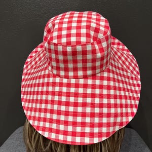 K&ouml;nnte beinhalten: Ein Sonnenhut mit breiter Krempe und rot-wei&szlig;em Vichy-Muster. Der Hut hat eine runde Krone und eine breite, kreisf&ouml;rmige Krempe, die viel Schatten spendet. Der Stoff hat eine klassische, sommerliche &Auml;sthetik.
