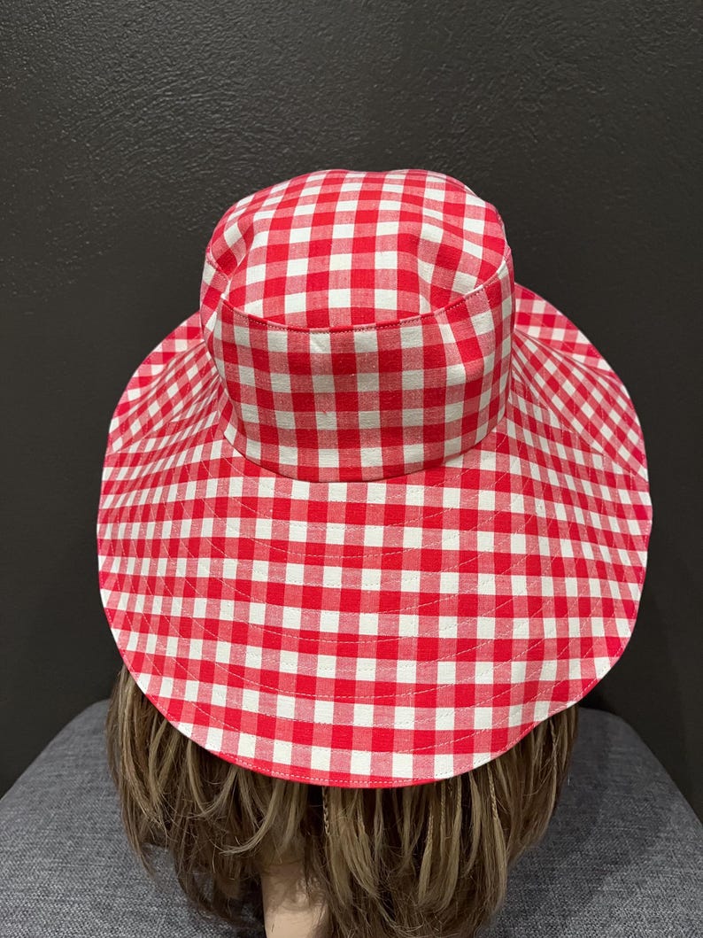 K&ouml;nnte beinhalten: Ein Sonnenhut mit breiter Krempe und rot-wei&szlig;em Vichy-Muster. Der Hut hat eine runde Krone und eine breite, kreisf&ouml;rmige Krempe, die viel Schatten spendet. Der Stoff hat eine klassische, sommerliche &Auml;sthetik.