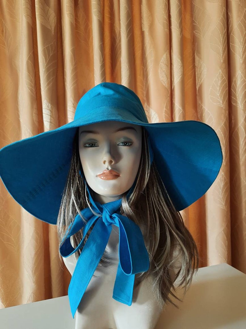 May include: A large, wide-brimmed sun hat in a bright turquoise blue. The hat has a wide brim and a simple design. The hat is tied with a bow at the neck.