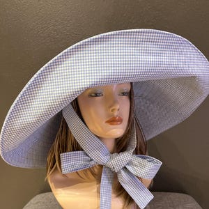 May include: A wide-brimmed hat with a blue and white gingham pattern. The hat features a large brim and a matching bow tied under the chin. The hat is displayed on a mannequin head.