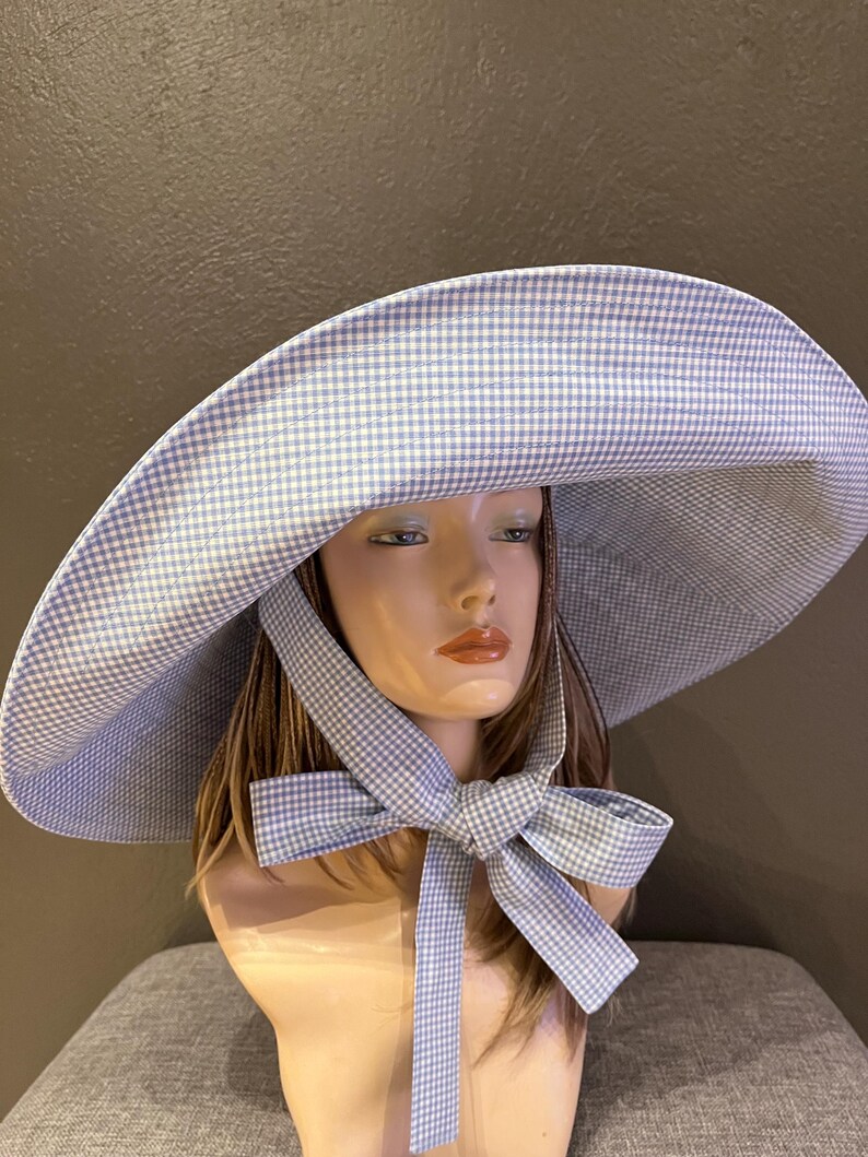 May include: A wide-brimmed hat with a blue and white gingham pattern. The hat features a large brim and a matching bow tied under the chin. The hat is displayed on a mannequin head.