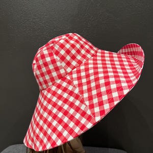 K&ouml;nnte beinhalten: Ein rot-wei&szlig; karierter Sonnenhut mit breiter Krempe. Der Hut ist aus Stoff gefertigt und hat ein klassisches Karomuster. Der Hut soll vor der Sonne sch&uuml;tzen.