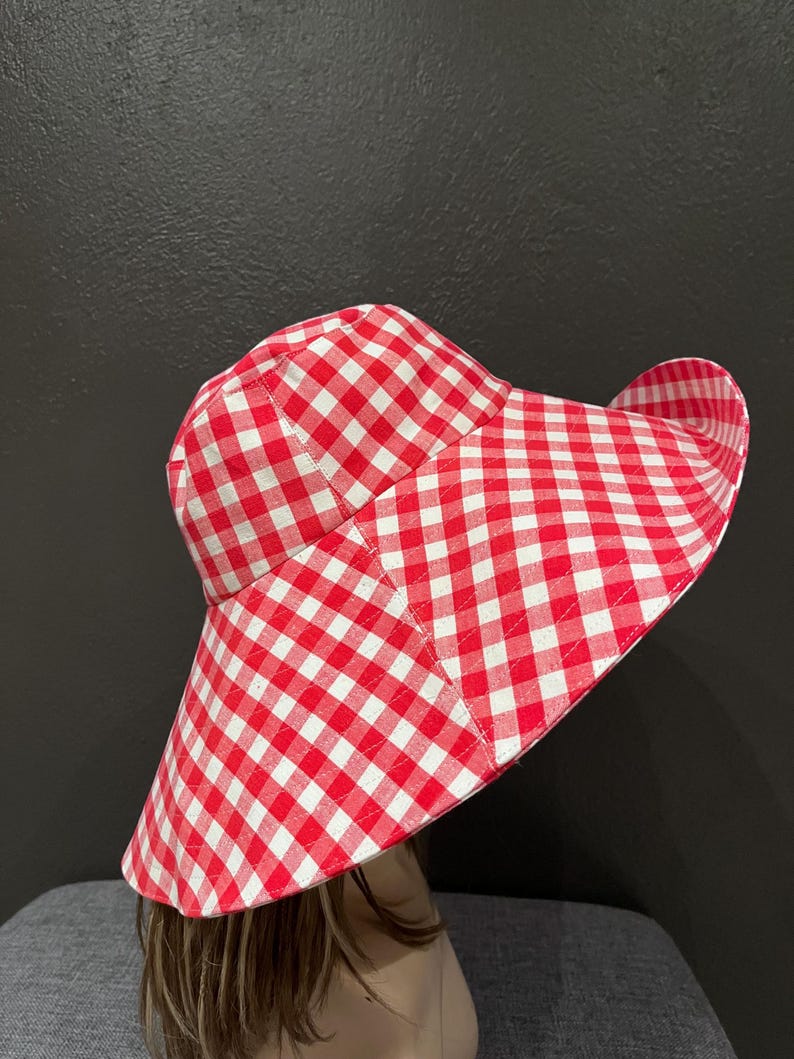 K&ouml;nnte beinhalten: Ein rot-wei&szlig; karierter Sonnenhut mit breiter Krempe. Der Hut ist aus Stoff gefertigt und hat ein klassisches Karomuster. Der Hut soll vor der Sonne sch&uuml;tzen.